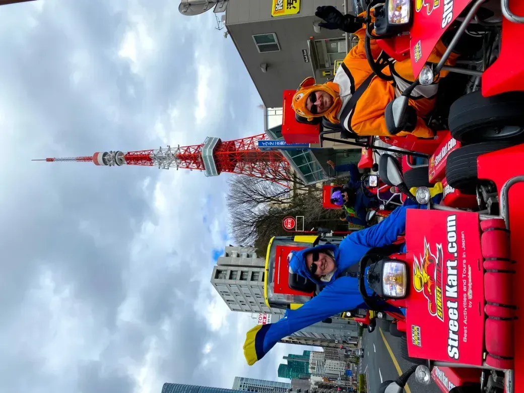 Group of people in bright costumes and helmets driving red go-karts along a city street, with a tall red-and-white tower in the background.