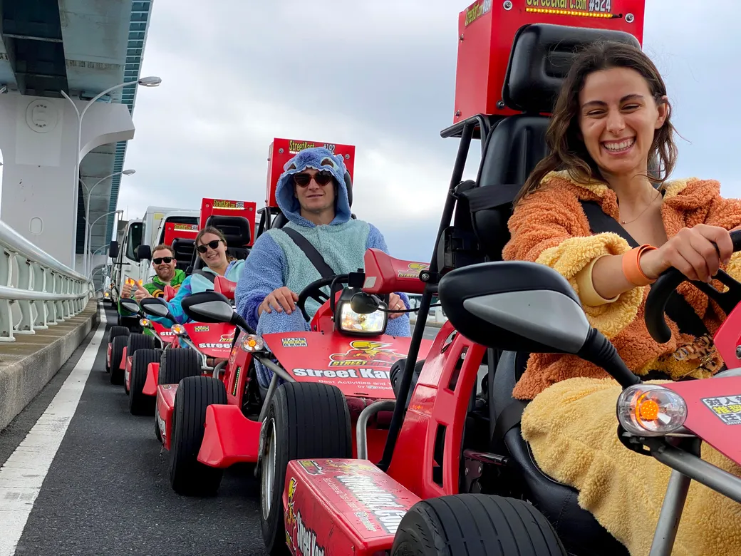 Group of friends riding red go-karts in a line on a roadway beside a bridge, smiling and wearing colorful costumes.